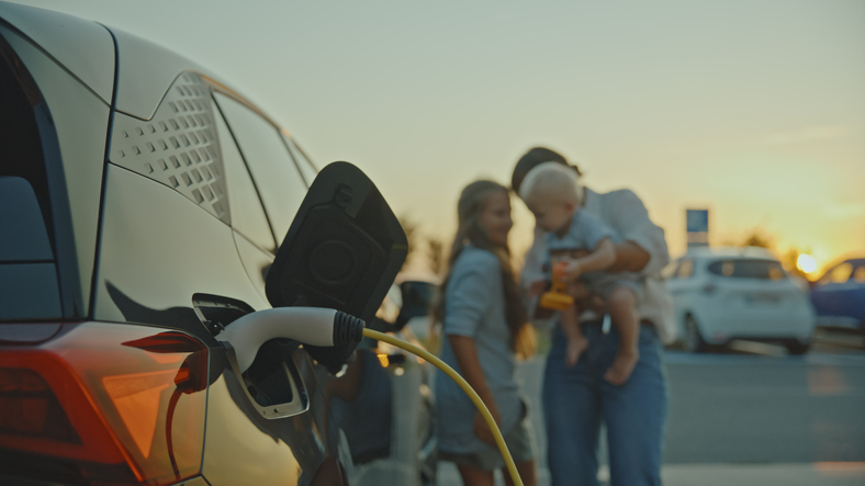 Caucasian Mother with Children Waiting Near Electric Car Charging at Sunset